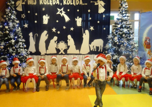 Chłopiec recytuje wiersz, trzyma w ręku mikrofon w tle dzieci siedzą na krzesłach na tle dekoracji świątecznej, choinek.